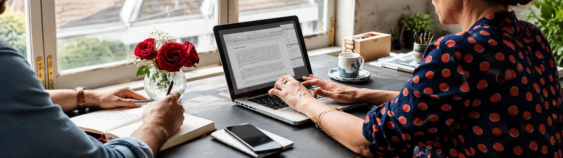 Two writers at a window table, one typing a document on a laptop and the other taking notes in a notebook