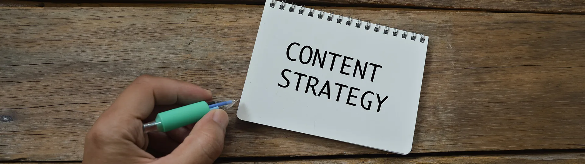 Hand holding a pen beside a spiral notepad reading Content Strategy on a wooden table