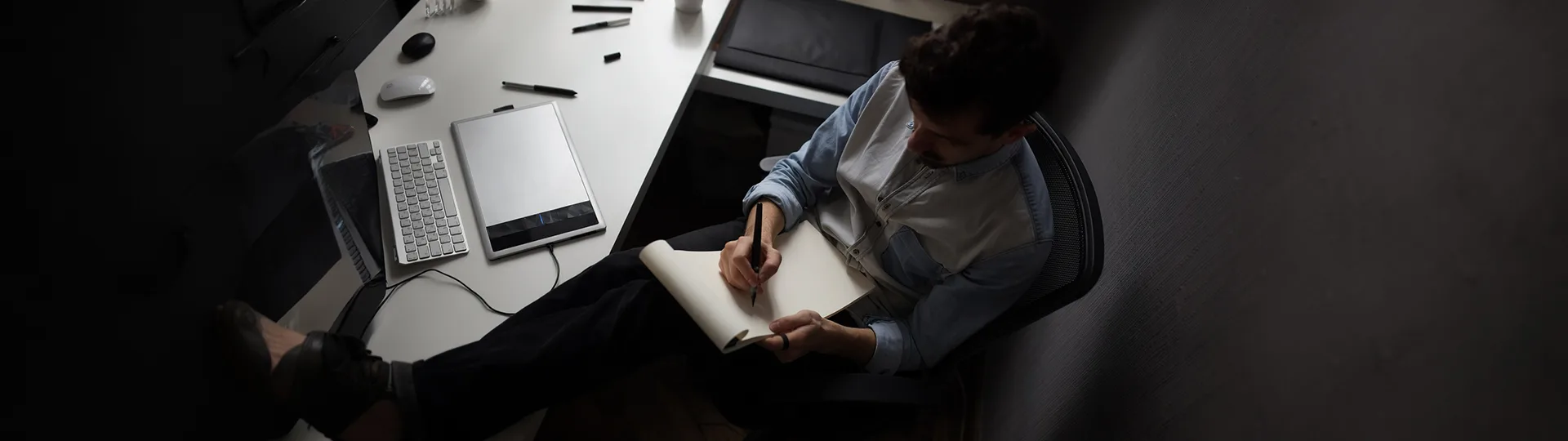Overhead view of a writer with his feet on a desk, writing by hand in a notebook beside a keyboard and tablet