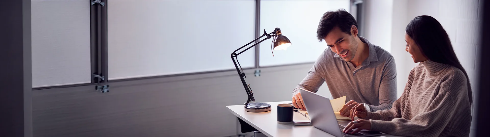 Two colleagues smiling while reviewing notes and a laptop at a desk lit by a lamp