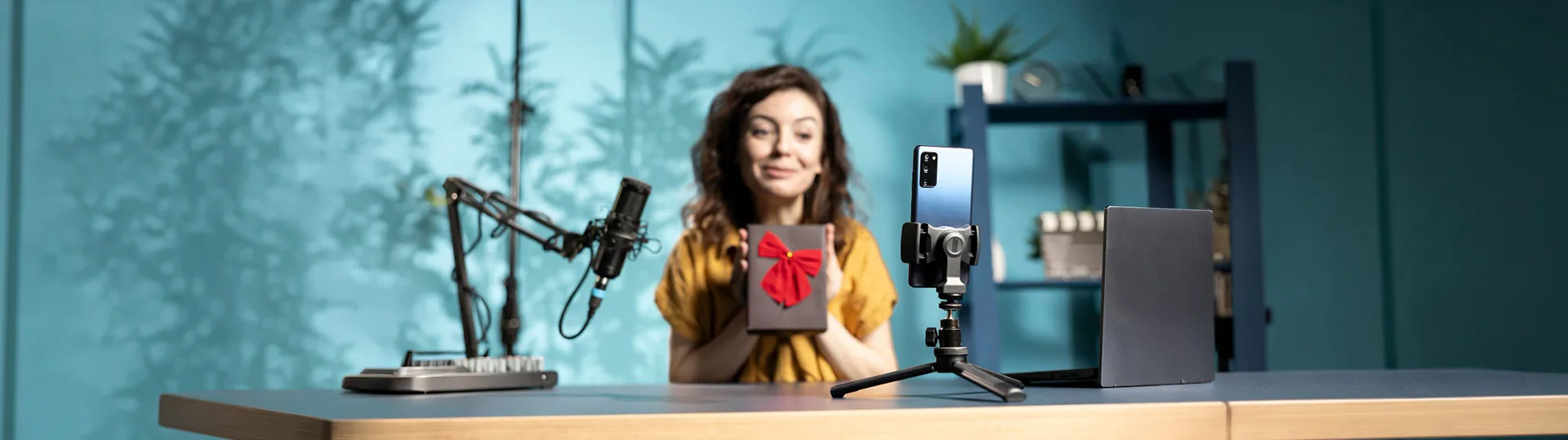 Creator holding up a gift box to a phone on a tripod, beside a podcast microphone and laptop