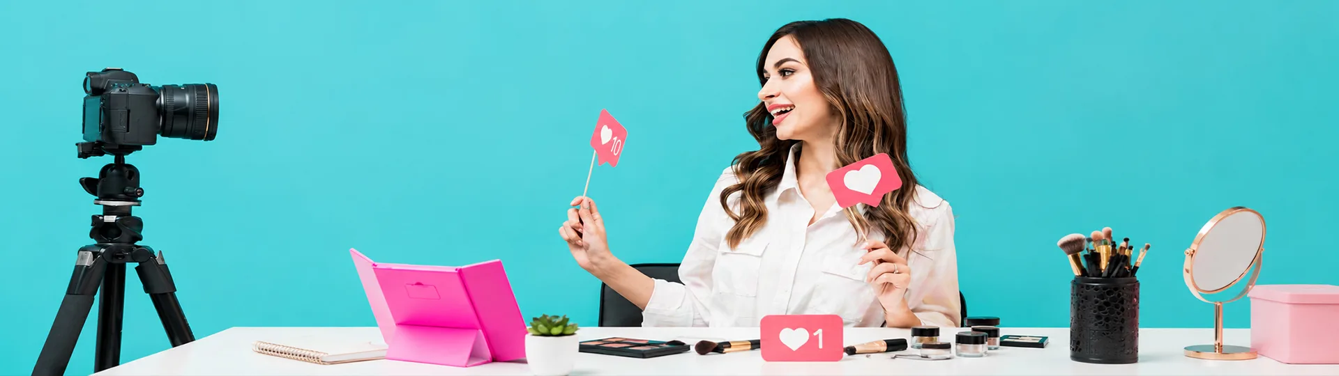 Beauty creator at a desk holding up like and heart signs while filming herself on a camera