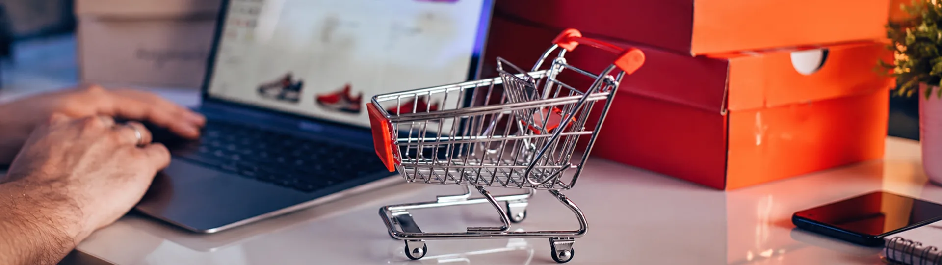 Miniature shopping cart beside a laptop showing an online store and red shoe boxes