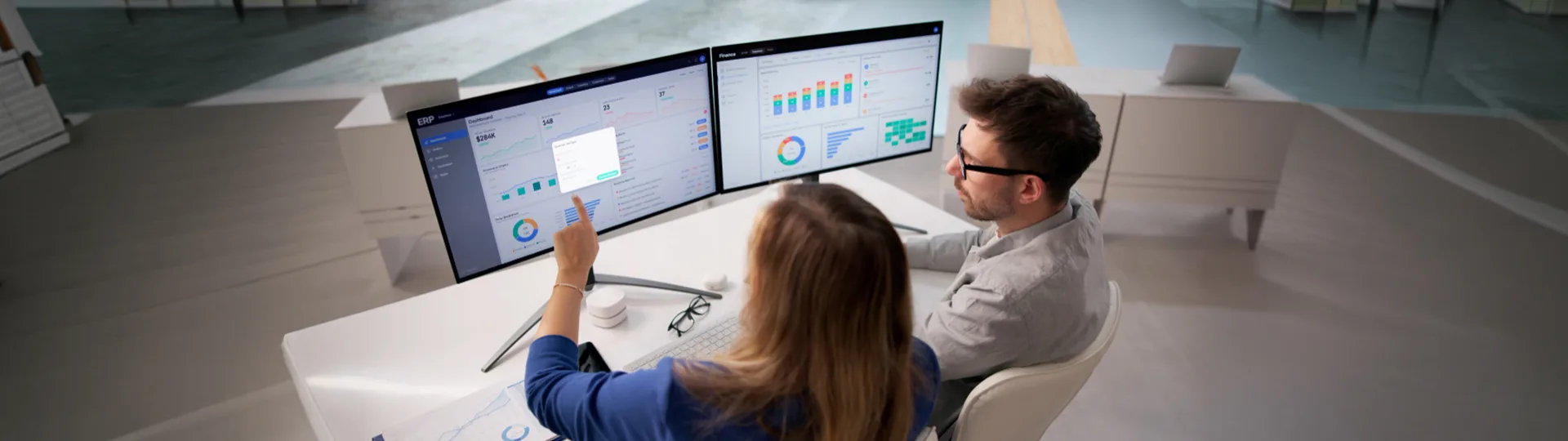 Two colleagues at a desk reviewing performance dashboards across two monitors