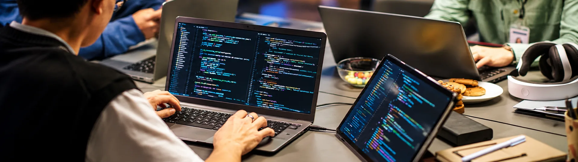 Developers coding on laptops and a tablet around a shared table, with code editors open on screen