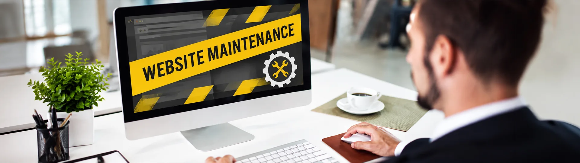 Man at a desk looking at a monitor showing a yellow Website Maintenance banner with a wrench and gear icon